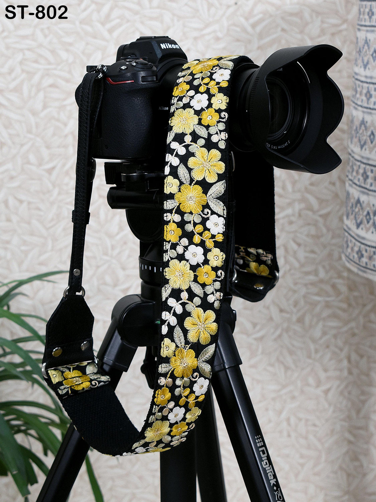 A black camera with a floral patterned strap is mounted on a tripod, with a potted plant visible in the background.