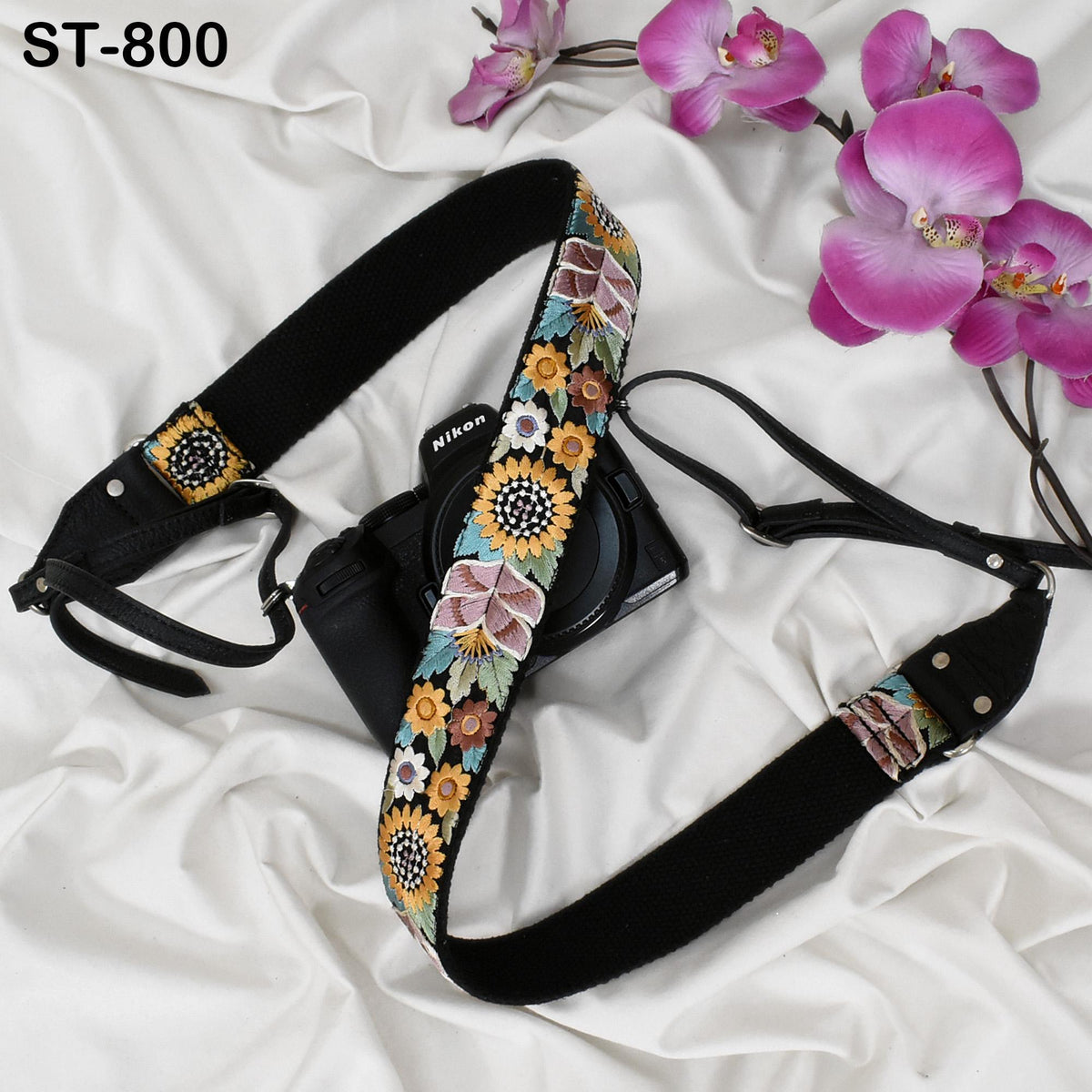 A black camera strap with a floral pattern is placed next to a camera on a white surface, with pink orchids in the background.