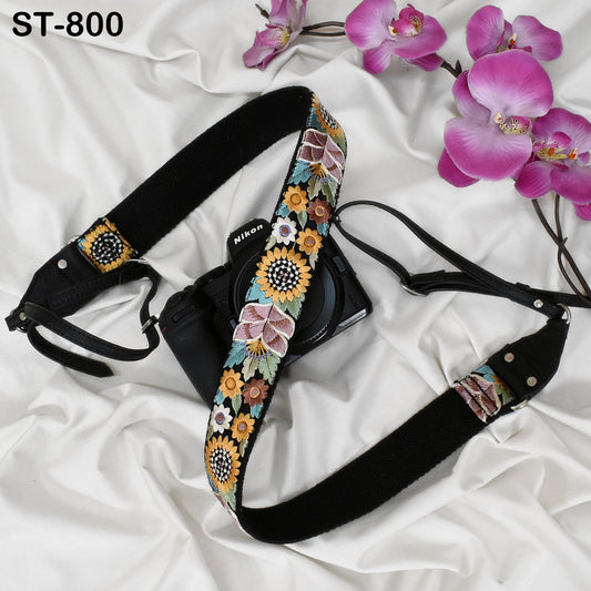 A black camera strap with a floral pattern is placed next to a camera on a white surface, with pink orchids in the background.