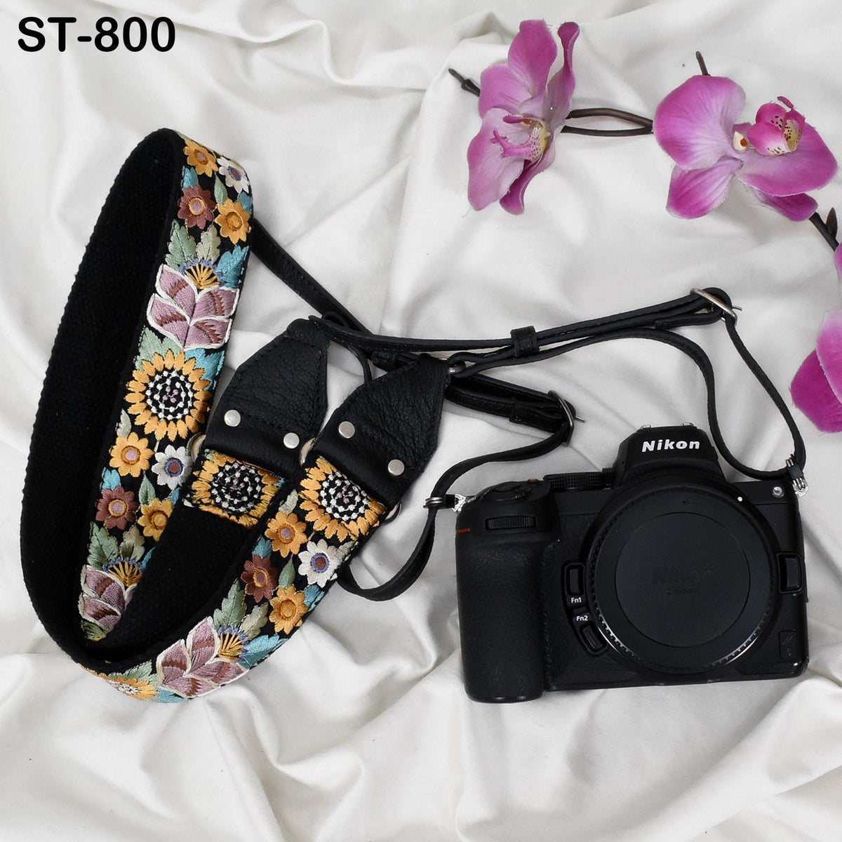 A black camera with a floral pattern strap and a pink orchid flower are placed on a white surface.