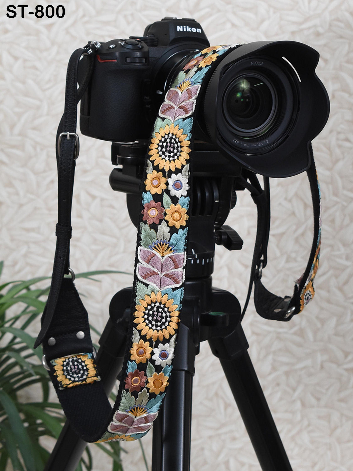 A black camera with a colorful floral patterned strap attached to it, placed on a tripod against a patterned background.