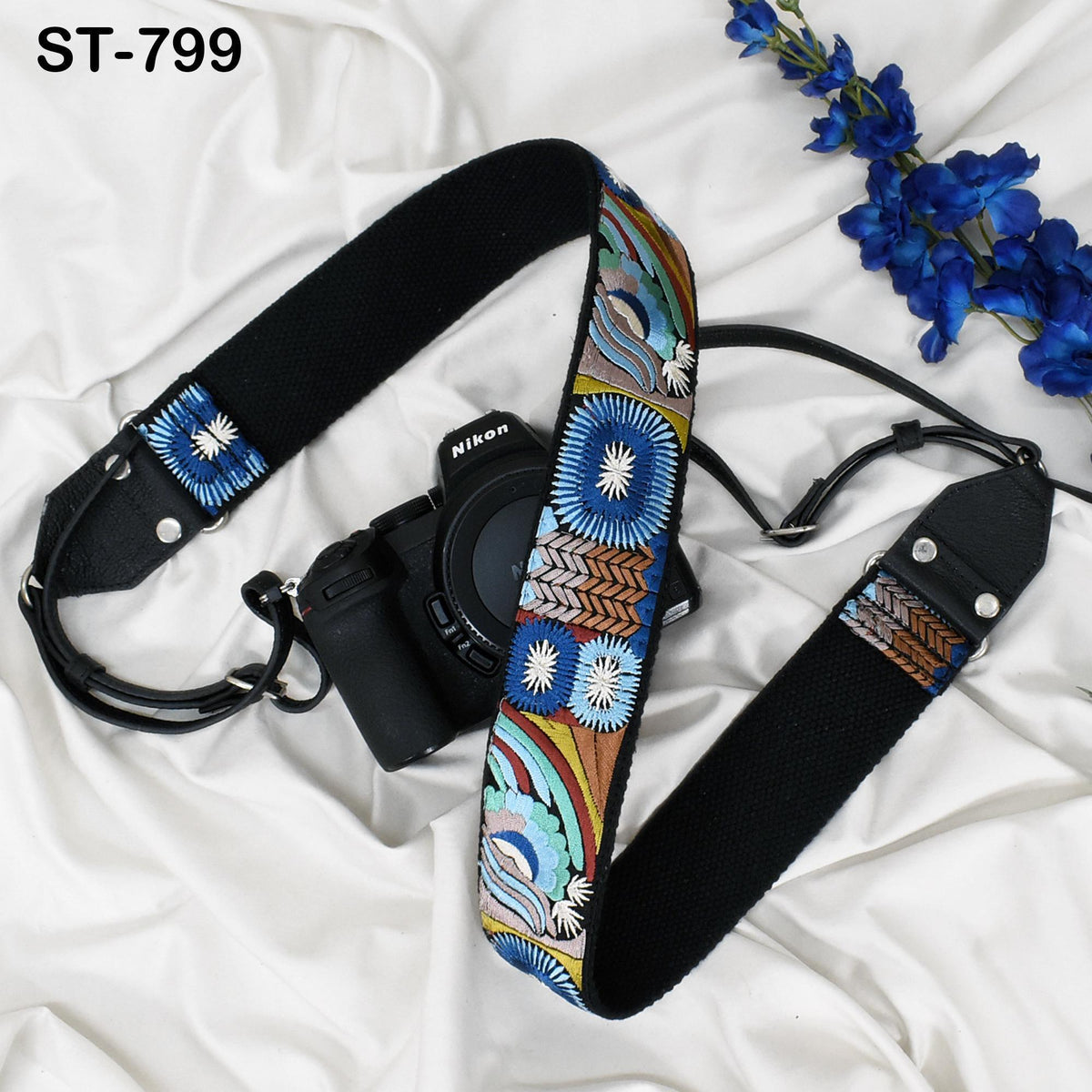 A black camera strap with a colorful, patterned design is placed next to a camera on a white surface, with a few blue flowers nearby.