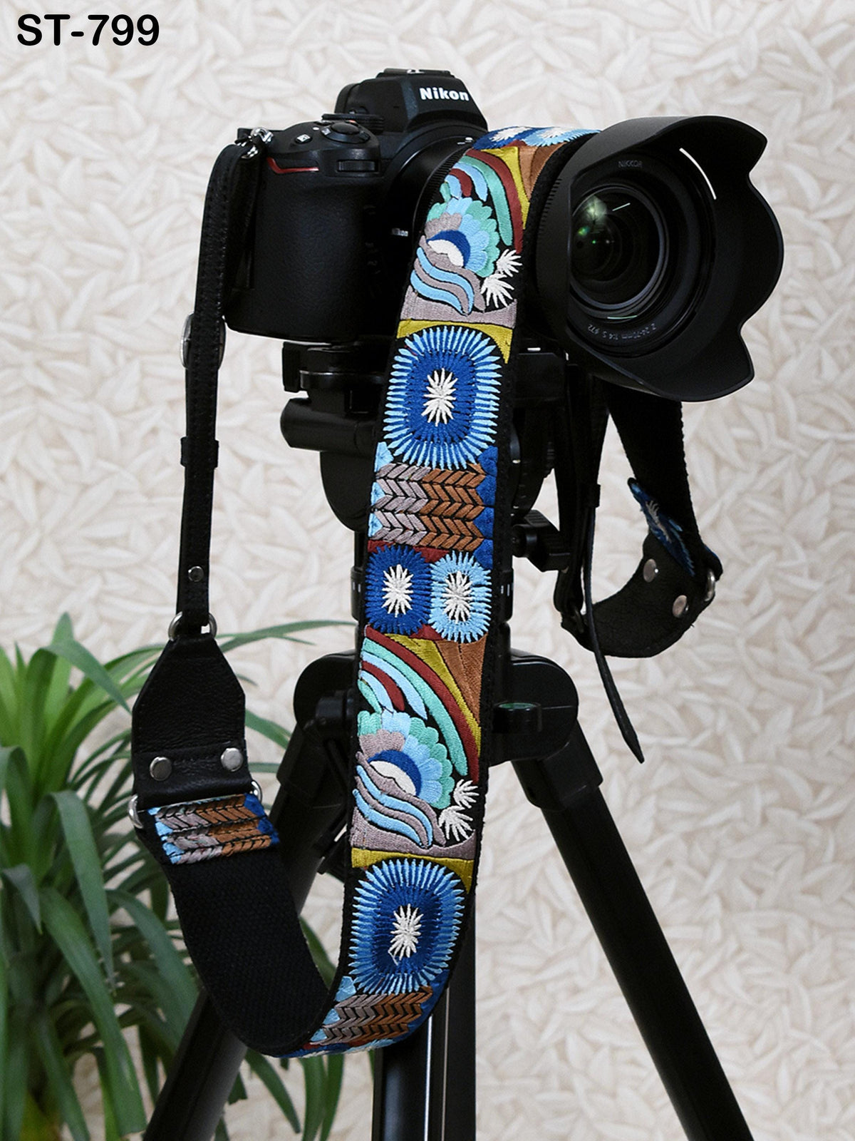 A black camera with a colorful patterned strap is mounted on a tripod, with a potted plant visible in the background.