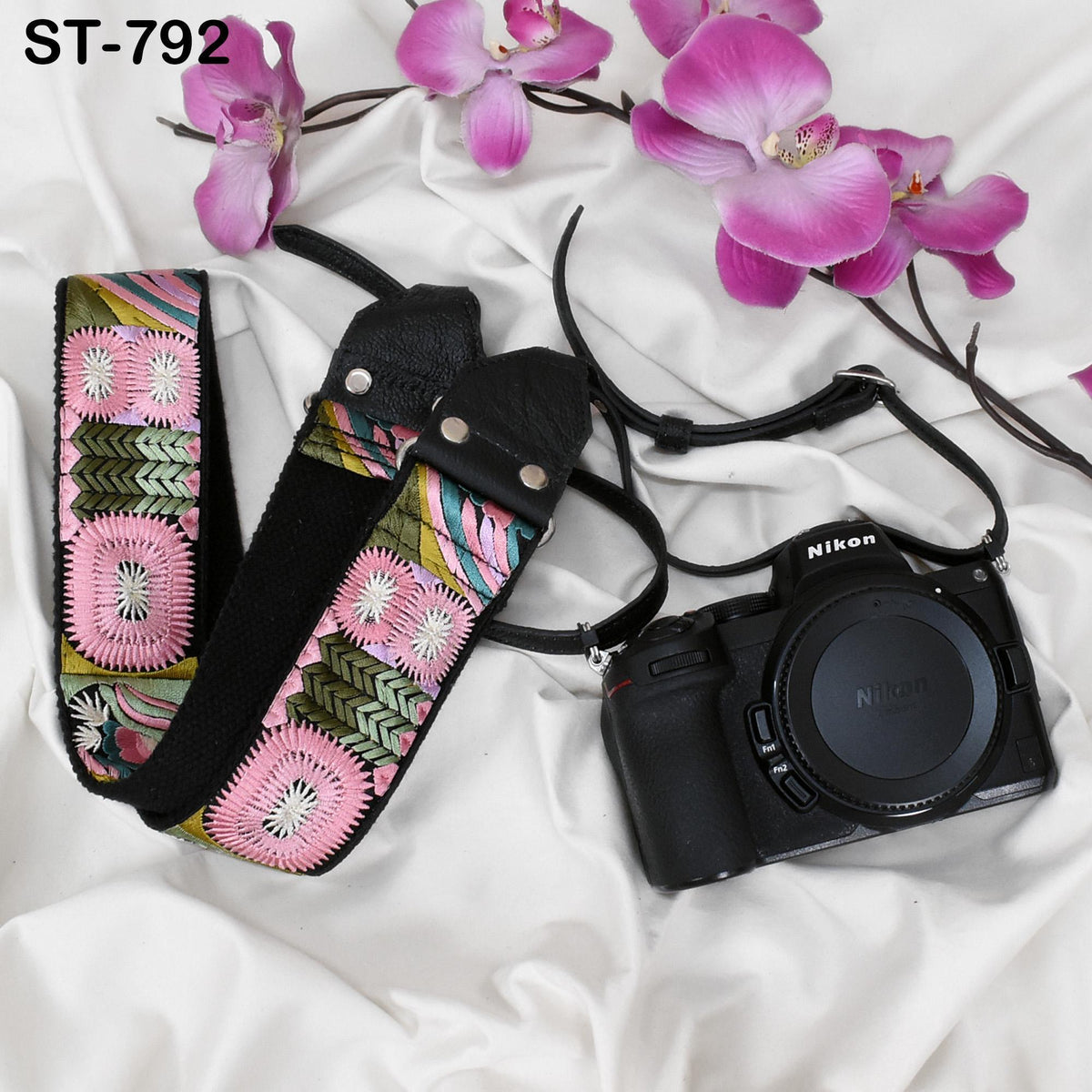 A black camera strap with a colorful floral pattern, a black camera, and pink orchids are arranged on a white background.