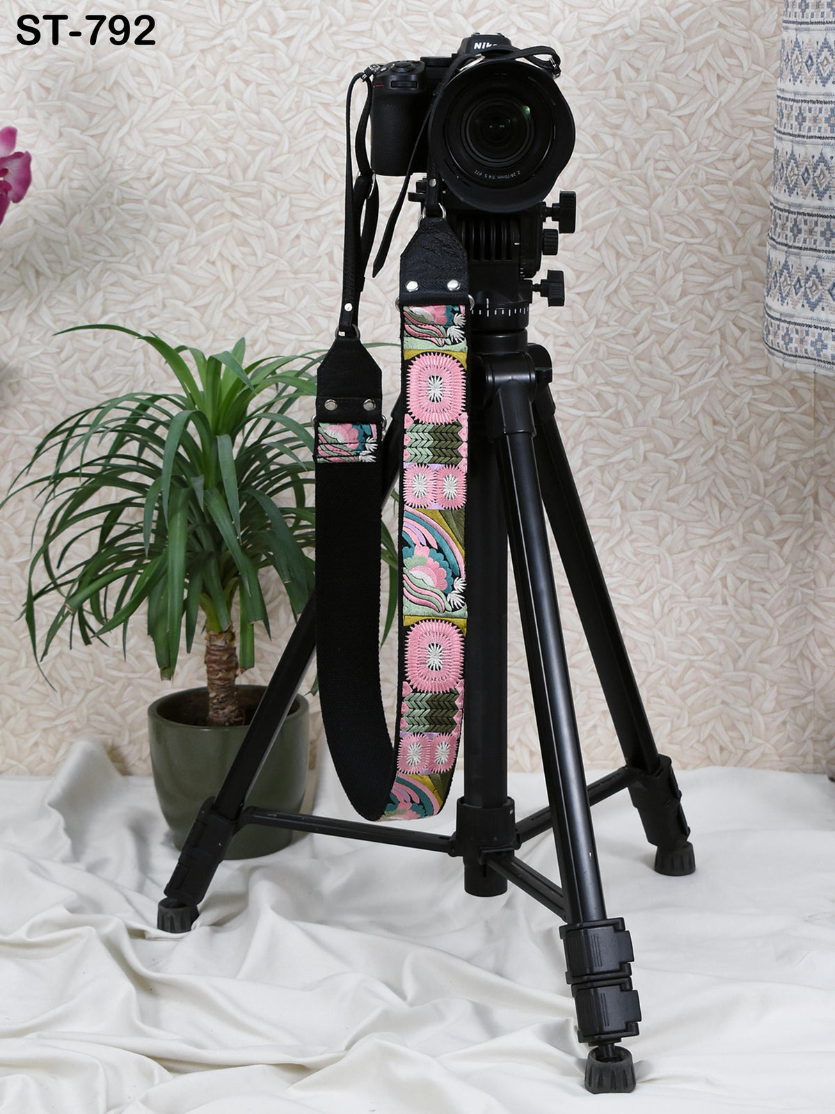 A black camera tripod with a colorful strap stands on a white surface, accompanied by a potted plant in the background.