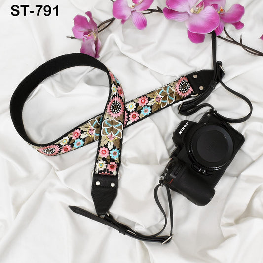 A floral patterned camera strap and a black camera with a lens attached are placed on a white fabric background, accompanied by pink orchids.