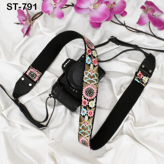 A black camera strap with a floral pattern and a black camera body are placed on a white surface, with pink orchids in the background.