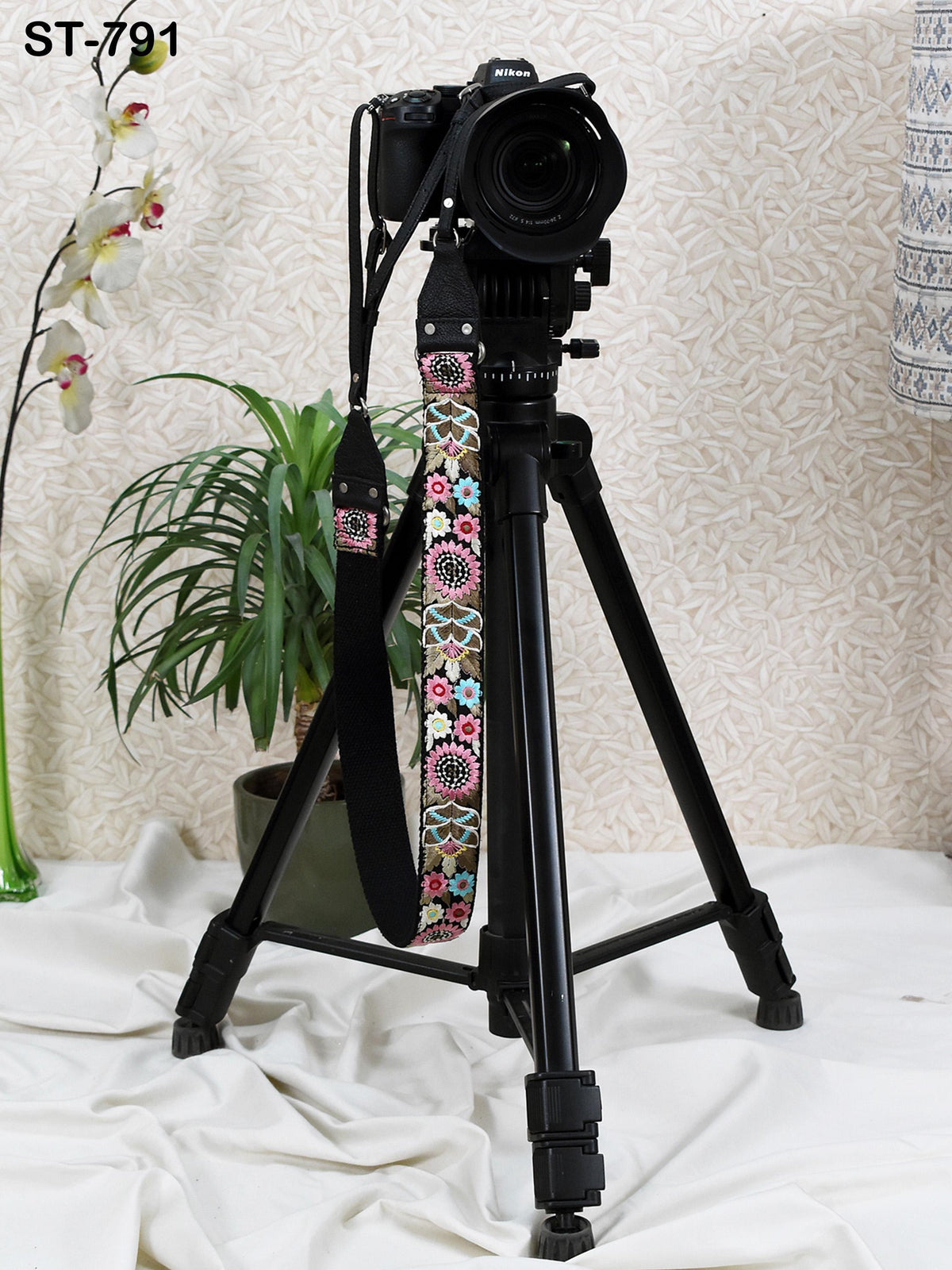 A black tripod with a camera attached to it stands on a white cloth, with a potted plant and a vase nearby.