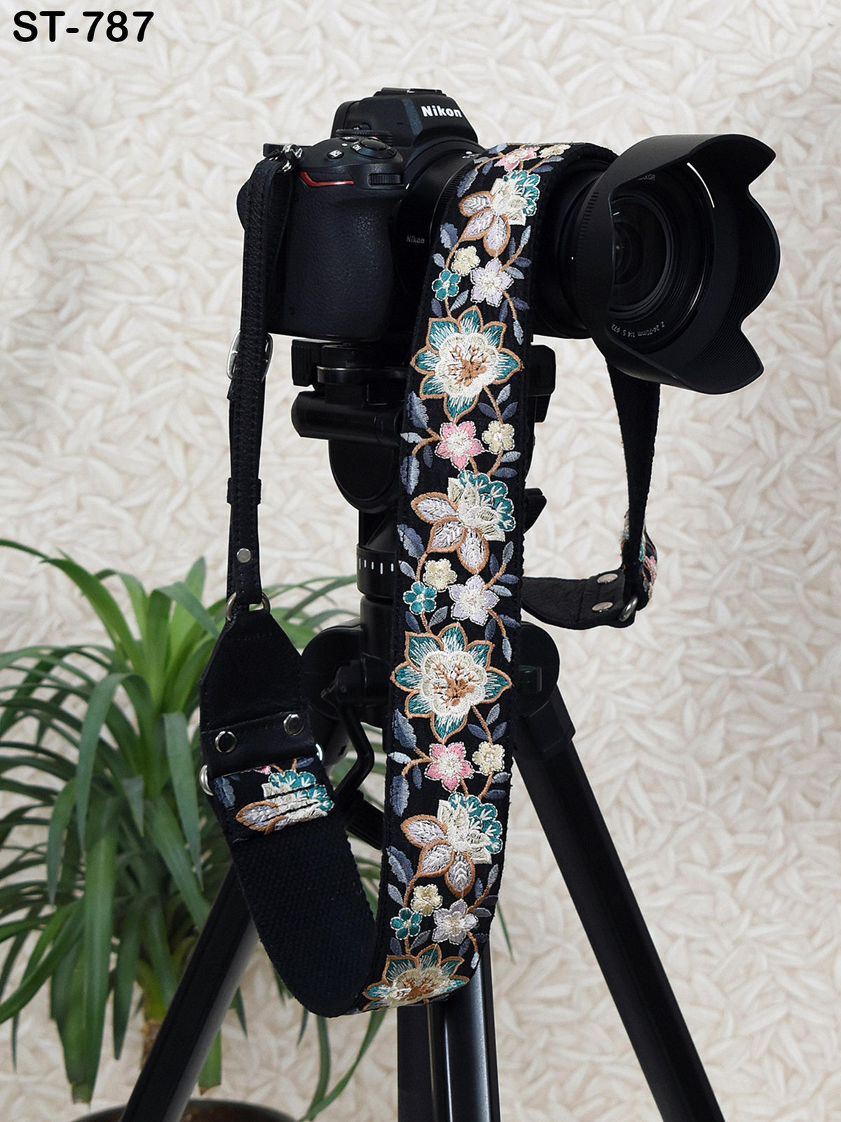 A black camera with a floral patterned strap is placed on a tripod, with a potted plant visible in the background.