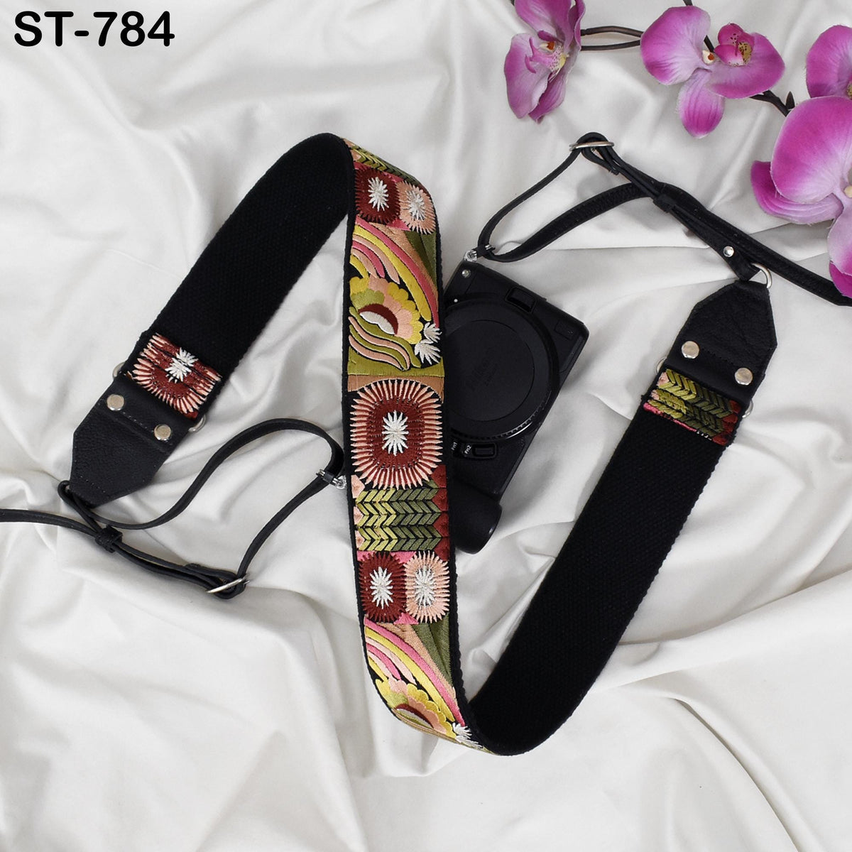 A camera strap with a colorful floral pattern is placed on a white surface, accompanied by a camera and some pink flowers.