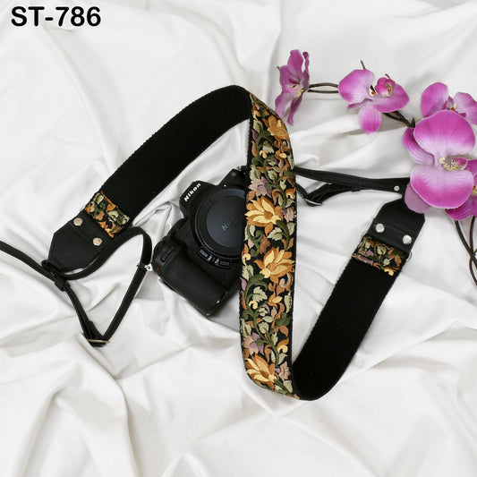 A black camera strap with a floral pattern and a black camera body are placed on a white surface, accompanied by a few pink orchid flowers.