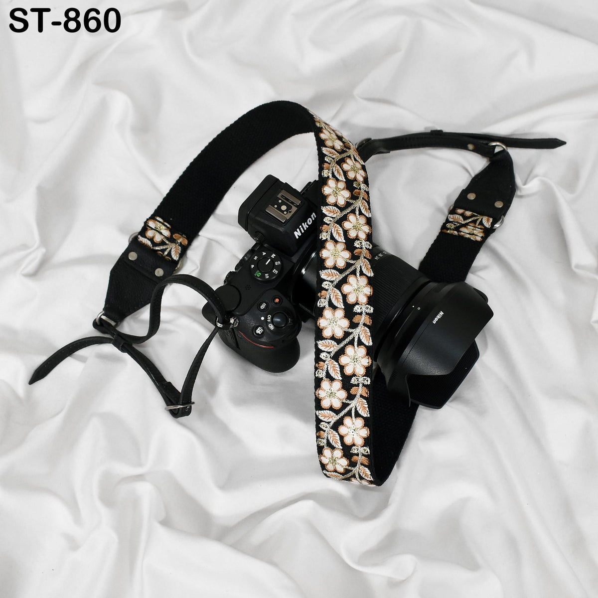 A black camera strap with a floral pattern and a camera attached to it, resting on a white fabric surface.