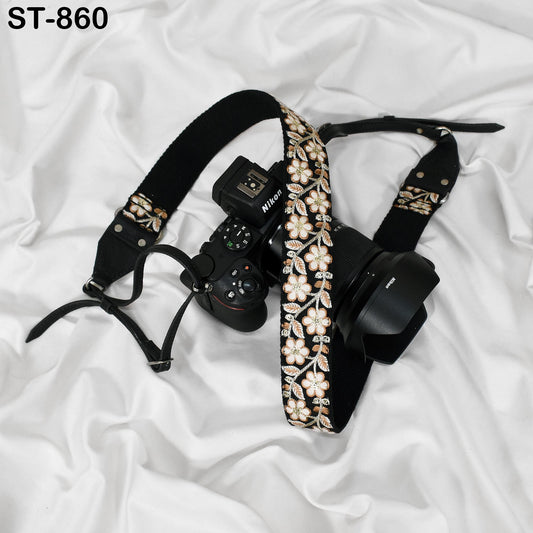 A black camera strap with a floral pattern and a camera attached to it, resting on a white fabric surface.