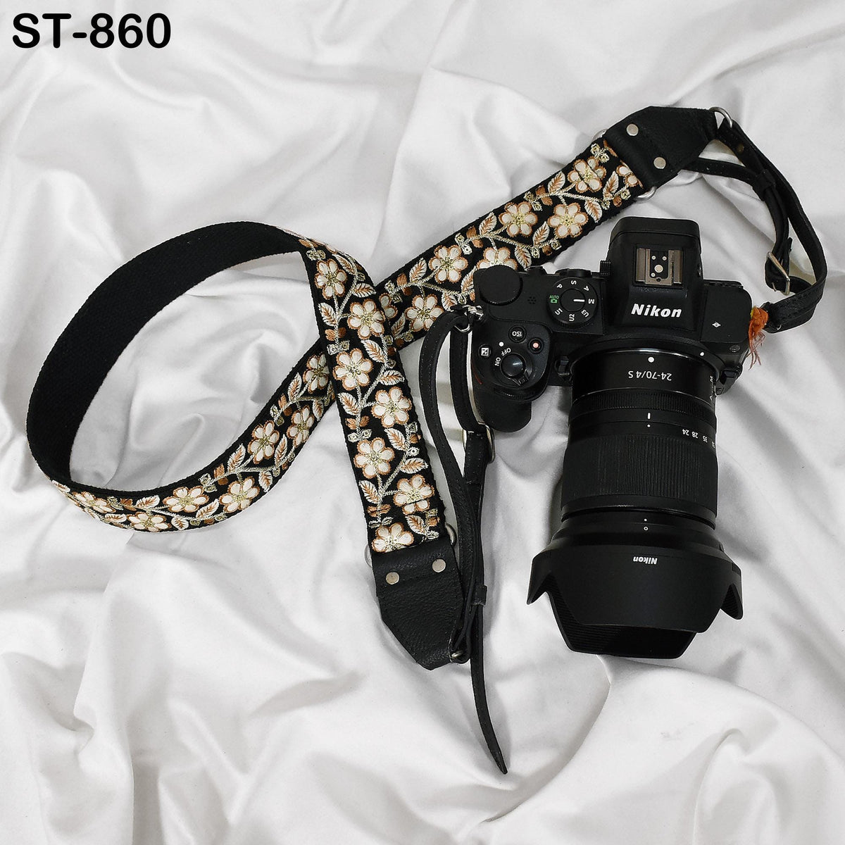 A black and gold floral camera strap and a Nikon camera are placed on a white fabric surface.