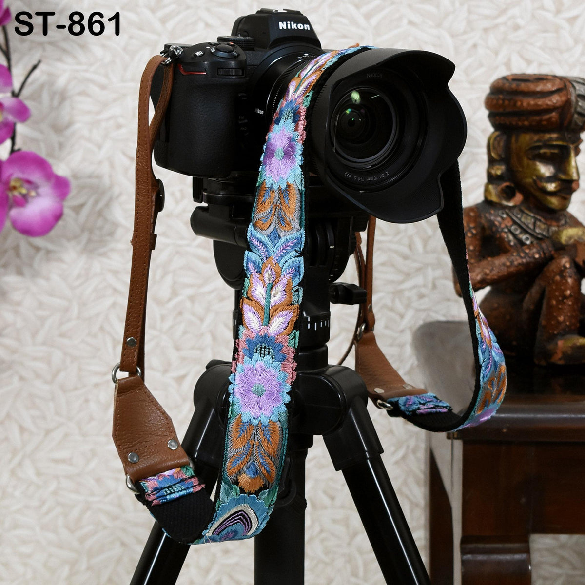 A black camera with a colorful floral strap is mounted on a tripod, with a decorative statue visible in the background.