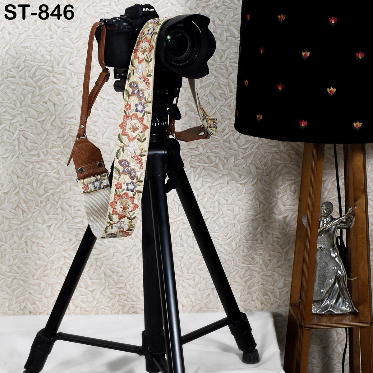A black tripod with a camera attached to it, along with a floral-patterned strap, is placed next to a lamp and a small statue on a table.