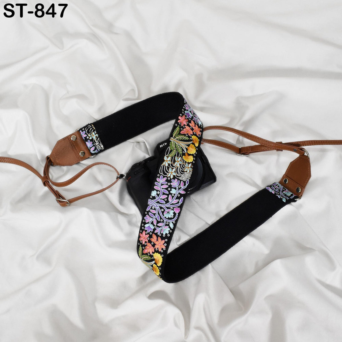 A black camera strap with a floral pattern and a brown leather strap are placed on a white fabric surface.
