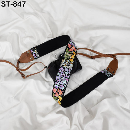 A black camera strap with a floral pattern and a brown leather strap are placed on a white fabric surface.