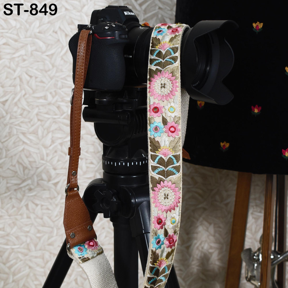 A black camera with a floral patterned strap is mounted on a tripod, with a black lens hood attached to the camera.