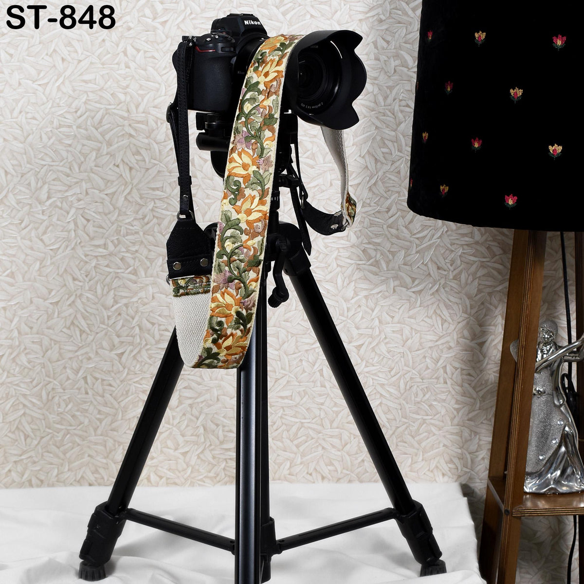 A black tripod with a camera attached to it, along with a colorful strap, is placed on a white surface.