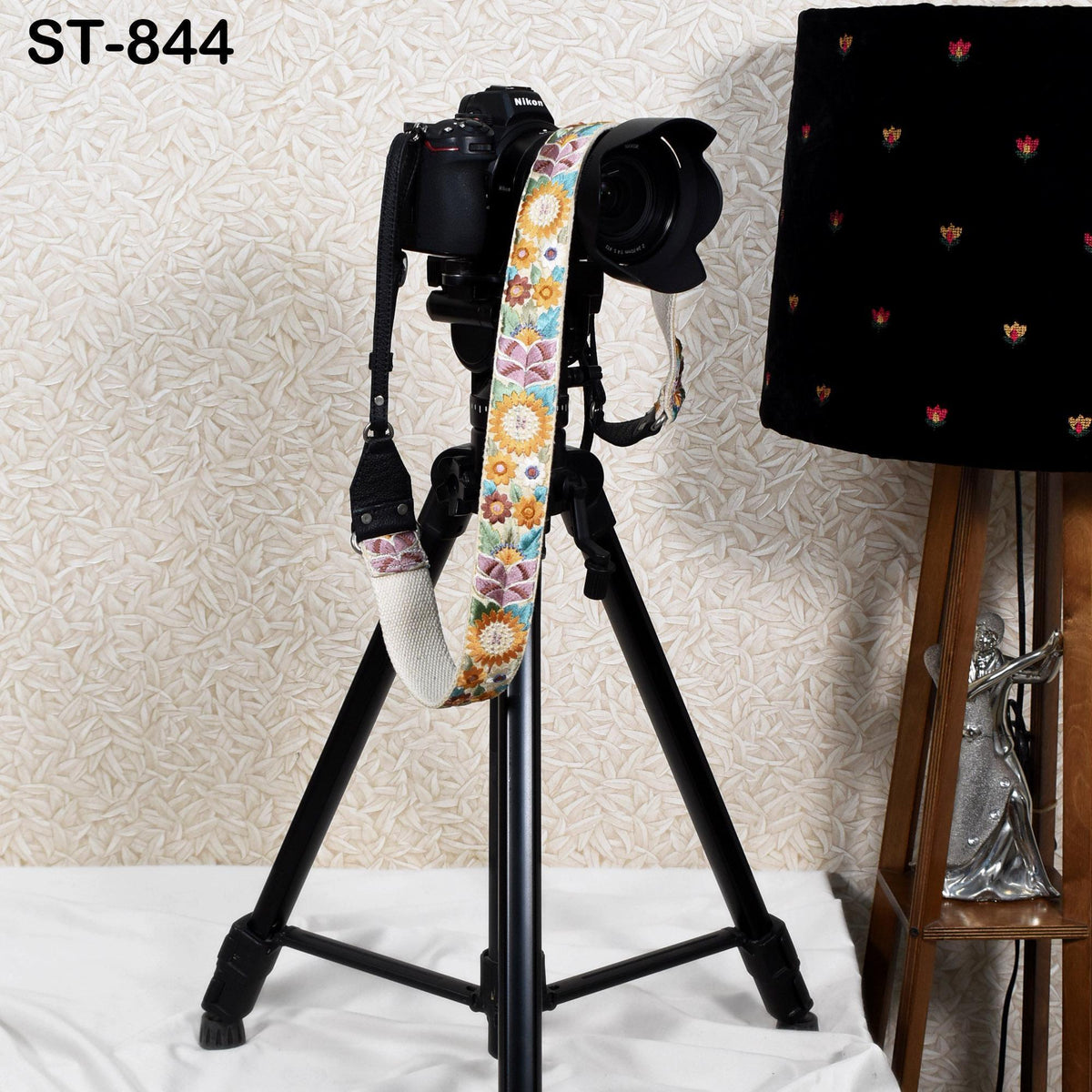 A black tripod with a colorful strap is placed on a white surface next to a black lamp with a floral pattern.