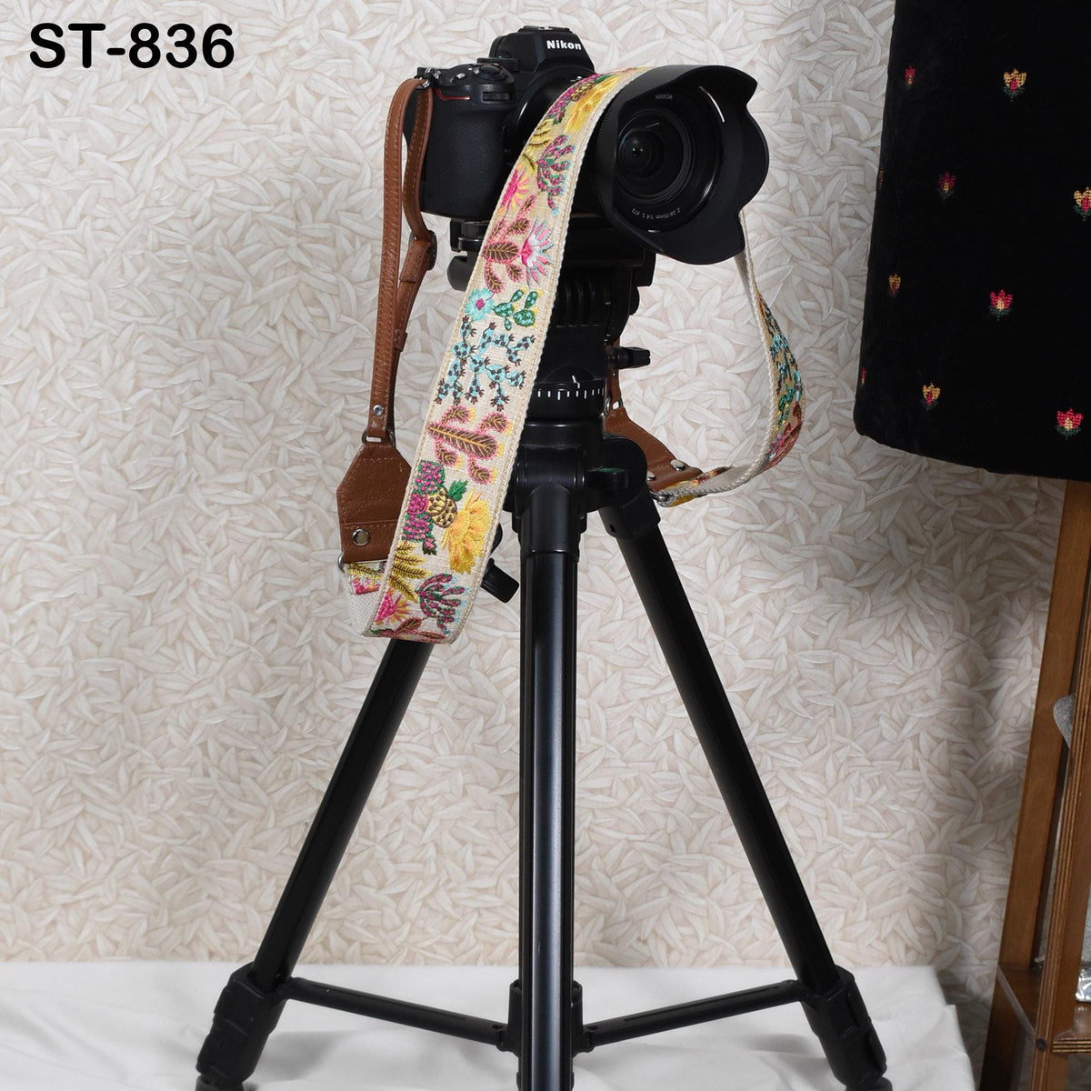 A black tripod with a colorful strap attached to it, standing on a white surface against a patterned wall.