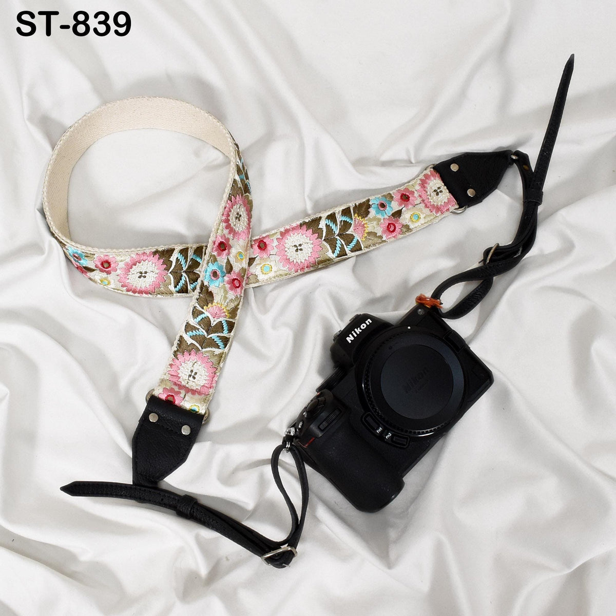 A floral patterned camera strap and a black camera with a lens attached are placed on a white fabric surface.