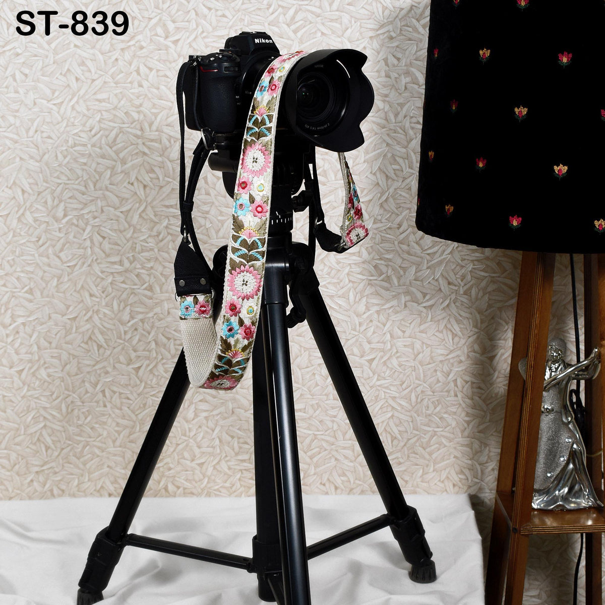 A black tripod with a camera attached to it, along with a floral patterned strap, is placed on a white surface next to a lamp.
