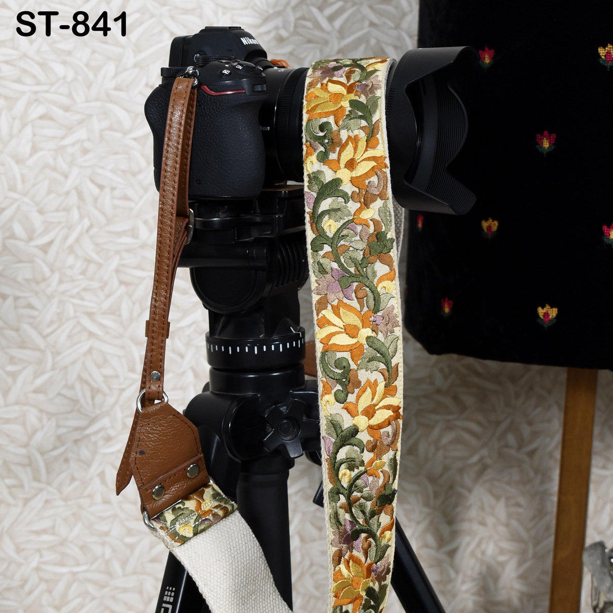 A black camera with a colorful floral patterned strap attached to it, placed on a tripod against a patterned background.