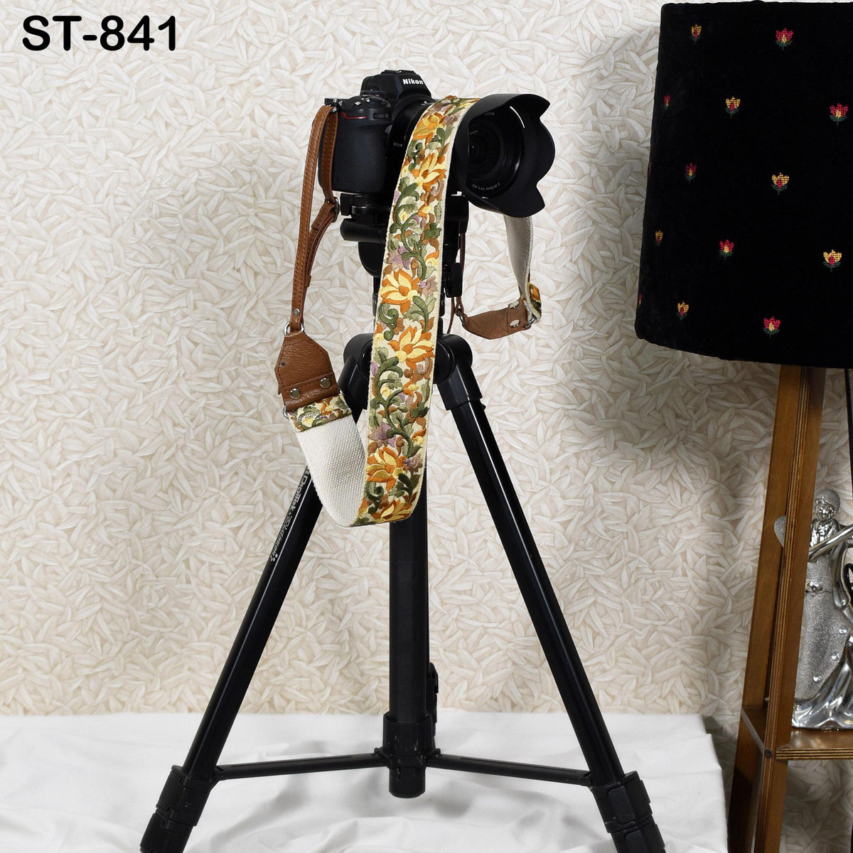 A black tripod with a camera strap attached to it, placed on a white surface against a patterned wall.