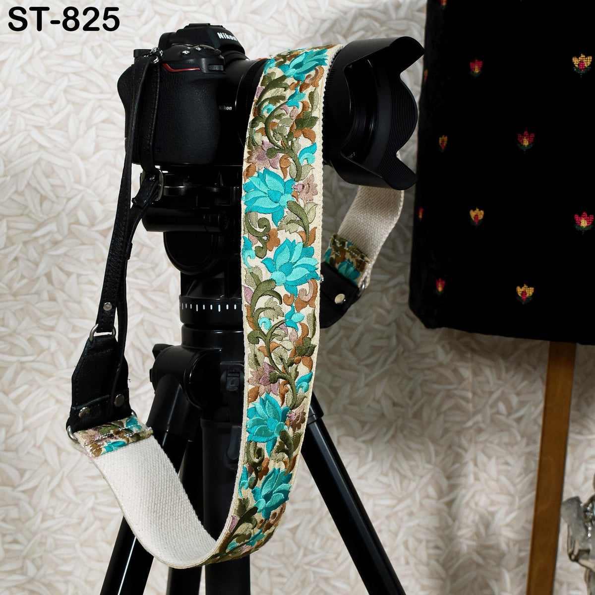 A black camera with a colorful floral patterned strap is placed on a tripod against a white background.