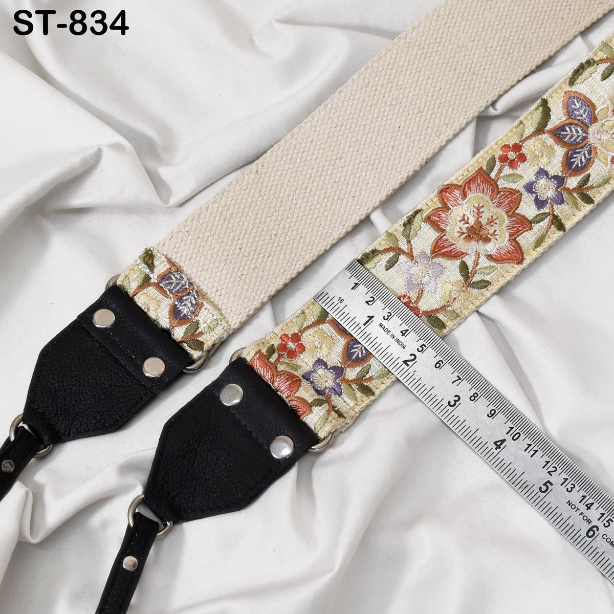 A beige strap with a floral pattern and a black leather strap with metal studs are placed on a white fabric surface. A ruler is visible, likely used for measuring the strap.