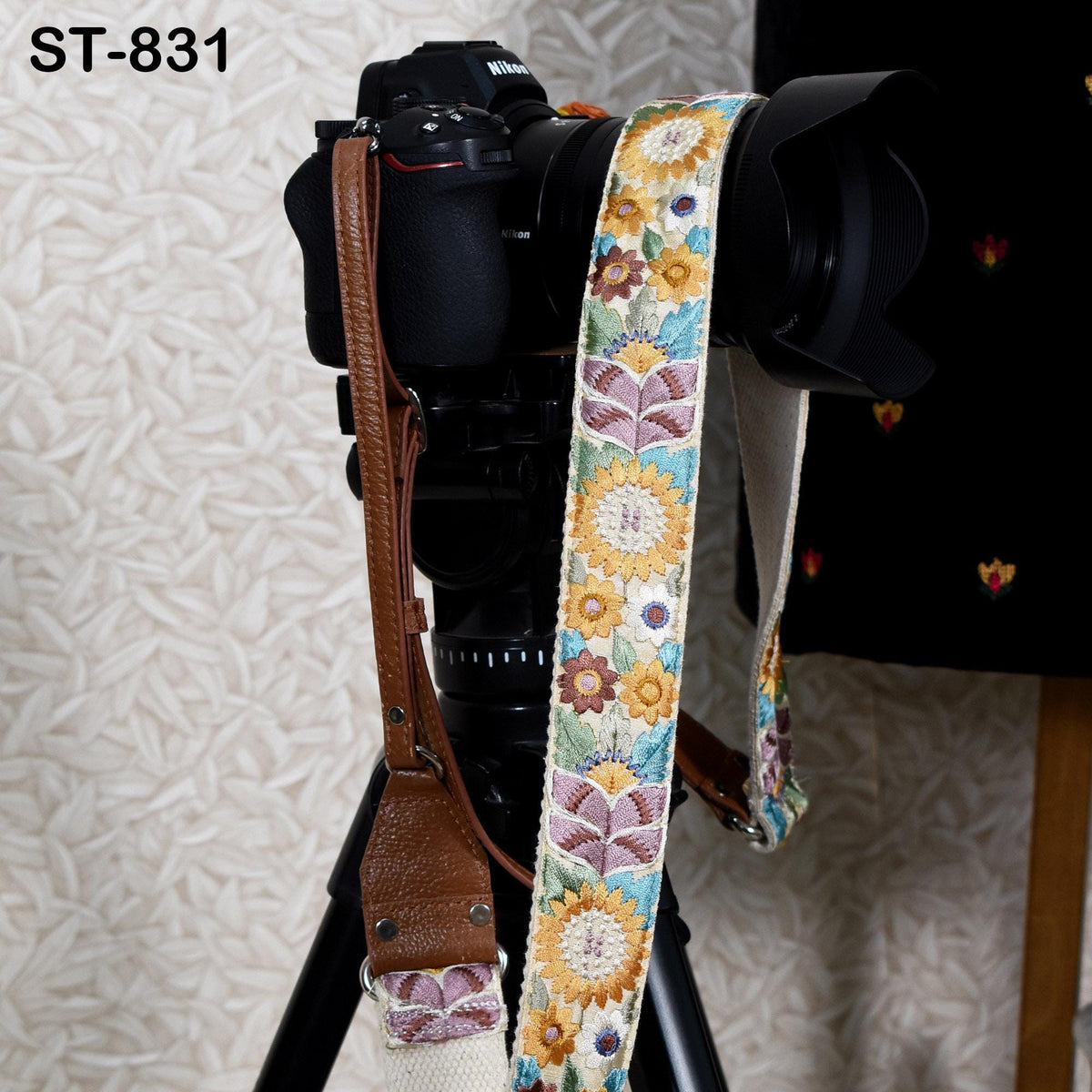 A black camera with a colorful floral strap hanging from it, placed on a tripod against a patterned background.
