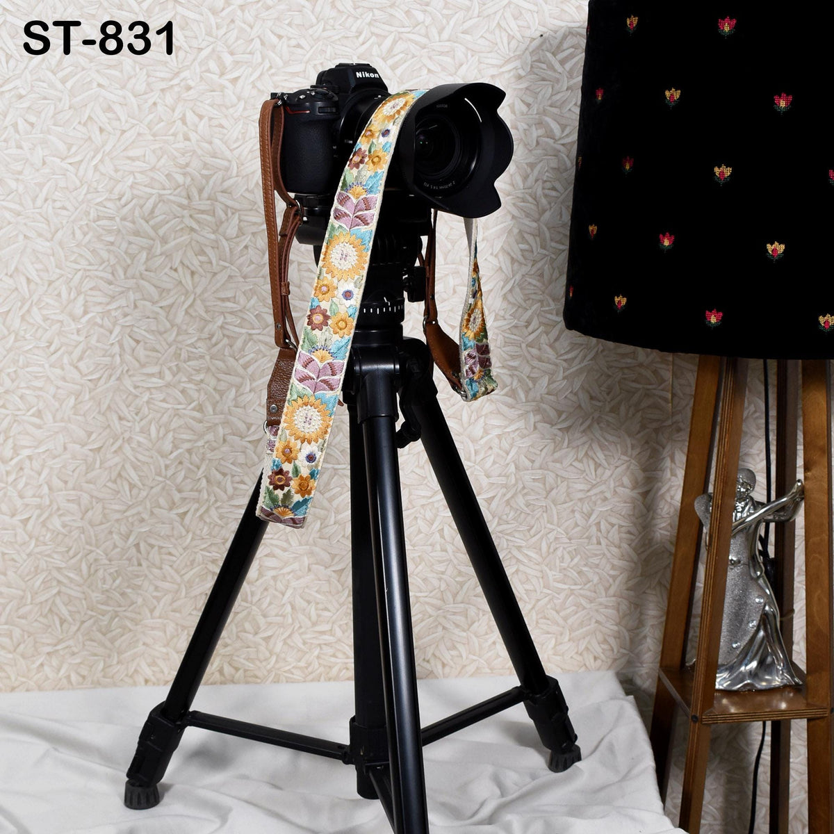 A black tripod with a camera strap and a colorful patterned strap is placed on a white surface, with a lamp and a figurine visible in the background.