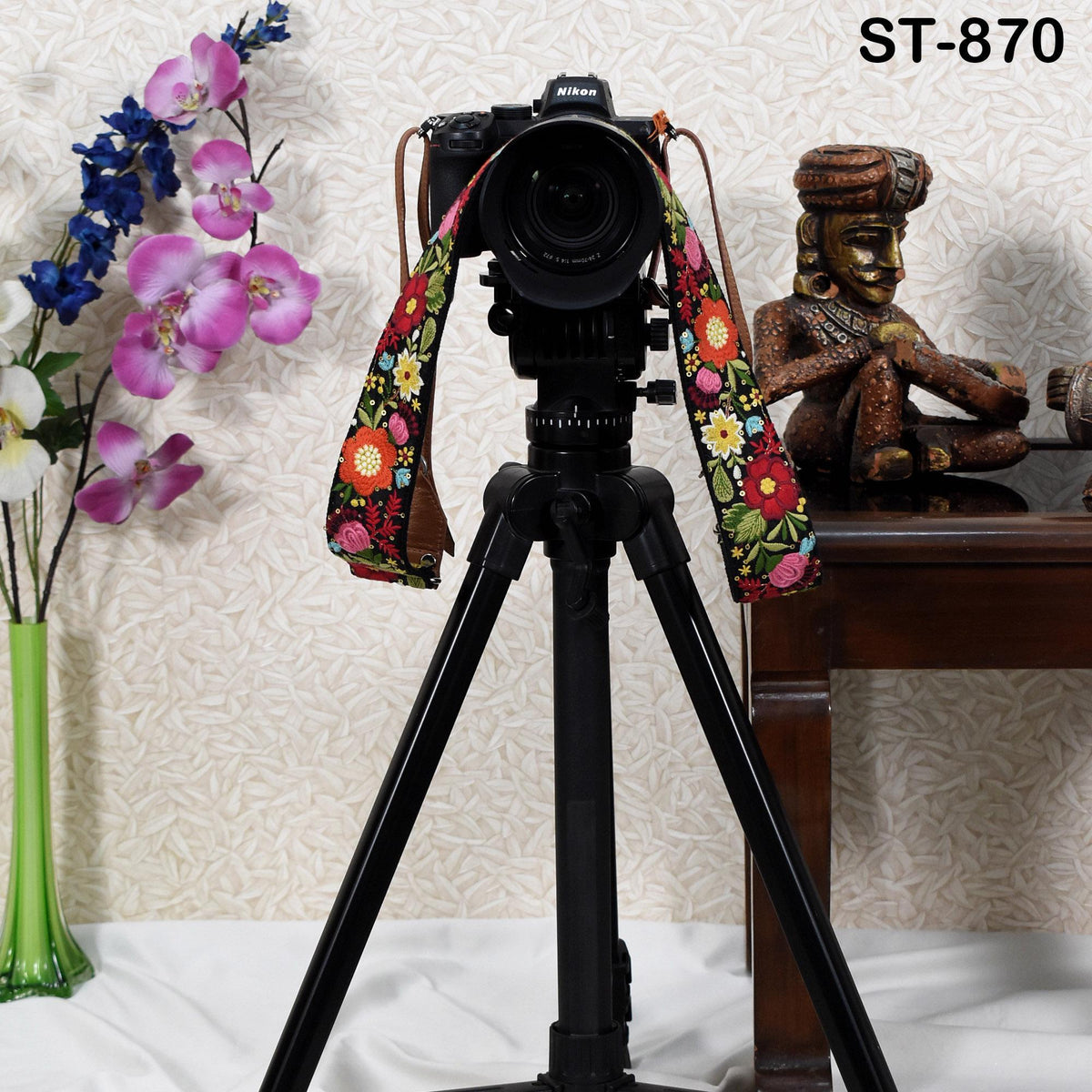A camera with a colorful strap is placed on a tripod, with a vase of flowers and a figurine nearby, against a patterned wall.
