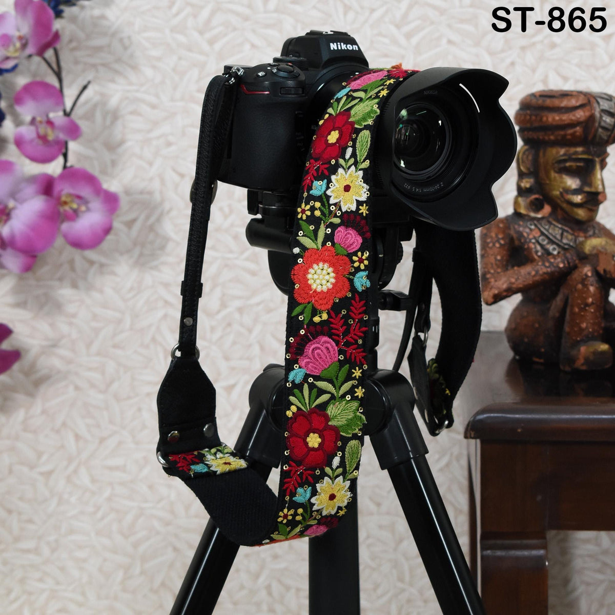 A black camera with a colorful floral strap is displayed on a tripod, with a small statue or figurine visible in the background.