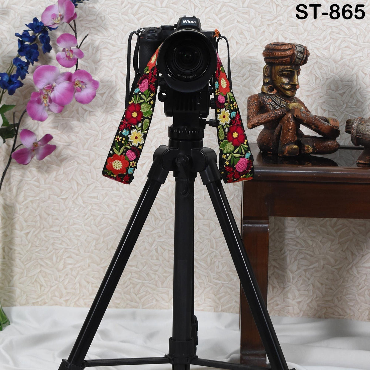 A black tripod with a colorful strap is placed on a wooden surface, with a small statue and a vase of flowers in the background.