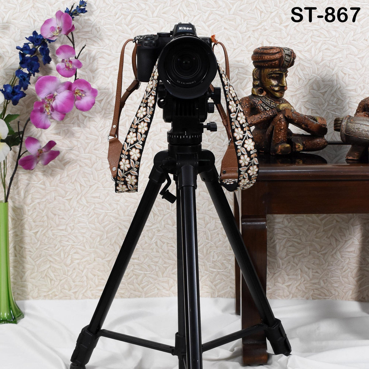 A black tripod with a camera attached to it, along with a strap with a floral pattern, is placed on a table next to a vase of purple flowers.