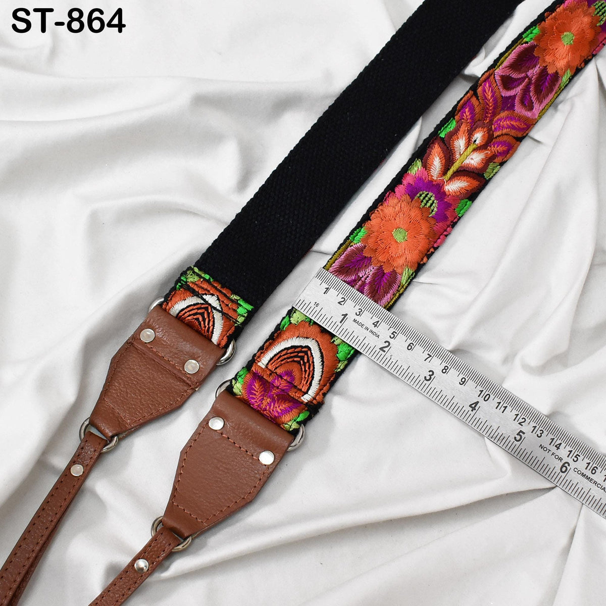 a colorful, patterned strap with a black leather buckle, placed on a white fabric surface. A ruler is visible in the foreground, indicating the strap's length.