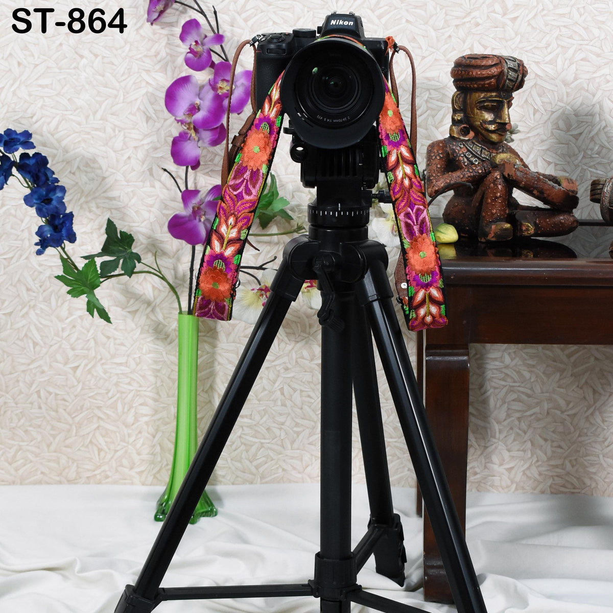 A black tripod with a camera attached to it, along with a colorful strap, is placed in front of a wooden table with a statue on it. The background features a floral arrangement and a wooden sculpture.