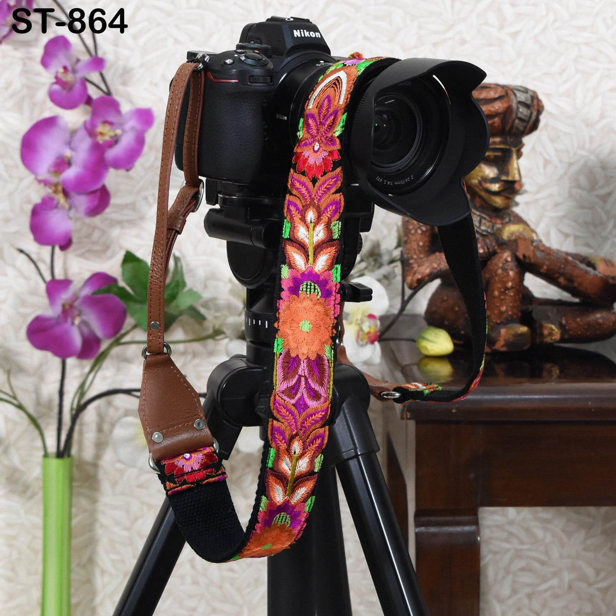 A black camera with a colorful, floral-patterned strap is mounted on a tripod, with a vase of purple orchids and a figurine in the background.