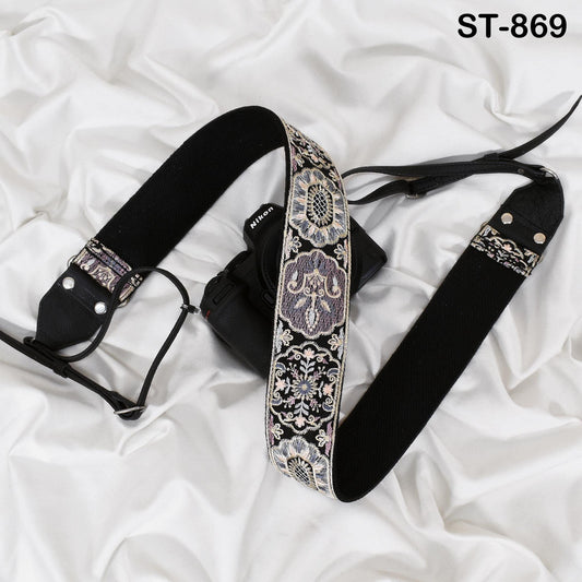 A black camera strap with a decorative pattern is placed on a white fabric surface.