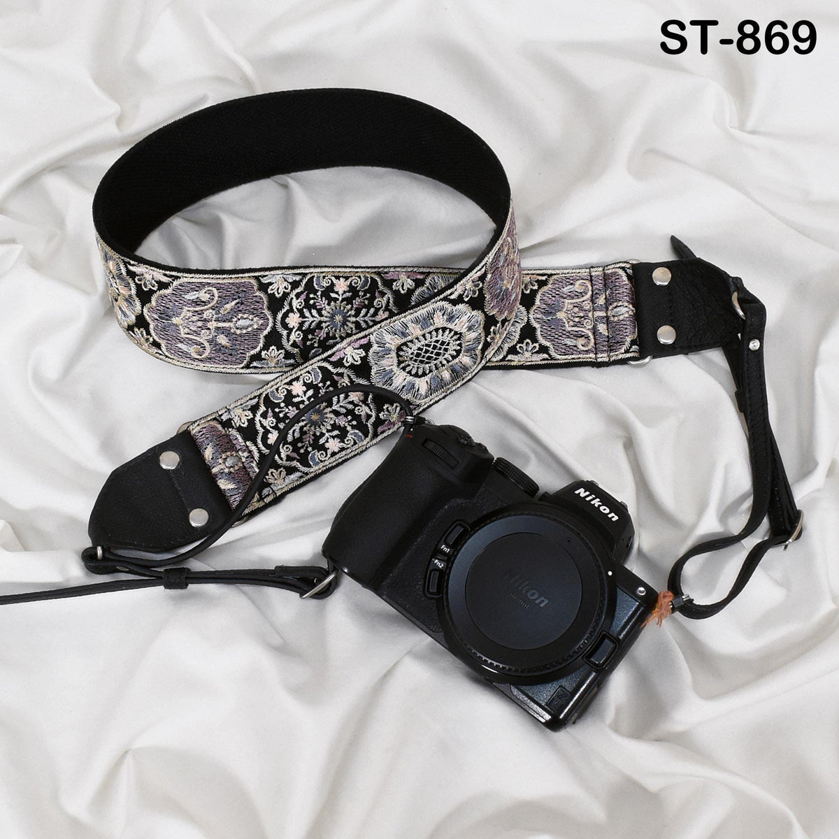A black and silver patterned camera strap and a black Nikon camera are placed on a white fabric surface.