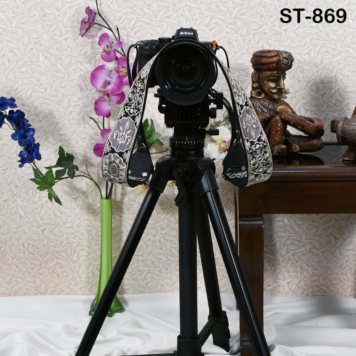 A black tripod with a camera attached to it, standing on a white surface. The tripod is positioned in front of a wooden table, which has a statue on it. The background features a wall with a floral pattern.