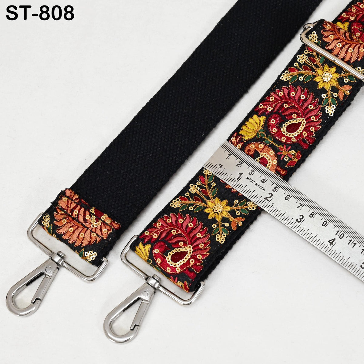 a black strap with a floral pattern and two metal hooks, placed next to a ruler for size comparison.