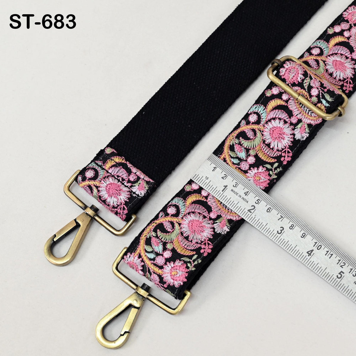 a black strap with a floral pattern and two gold-colored metal hooks attached to it. A ruler is placed next to the strap, likely for measuring purposes.