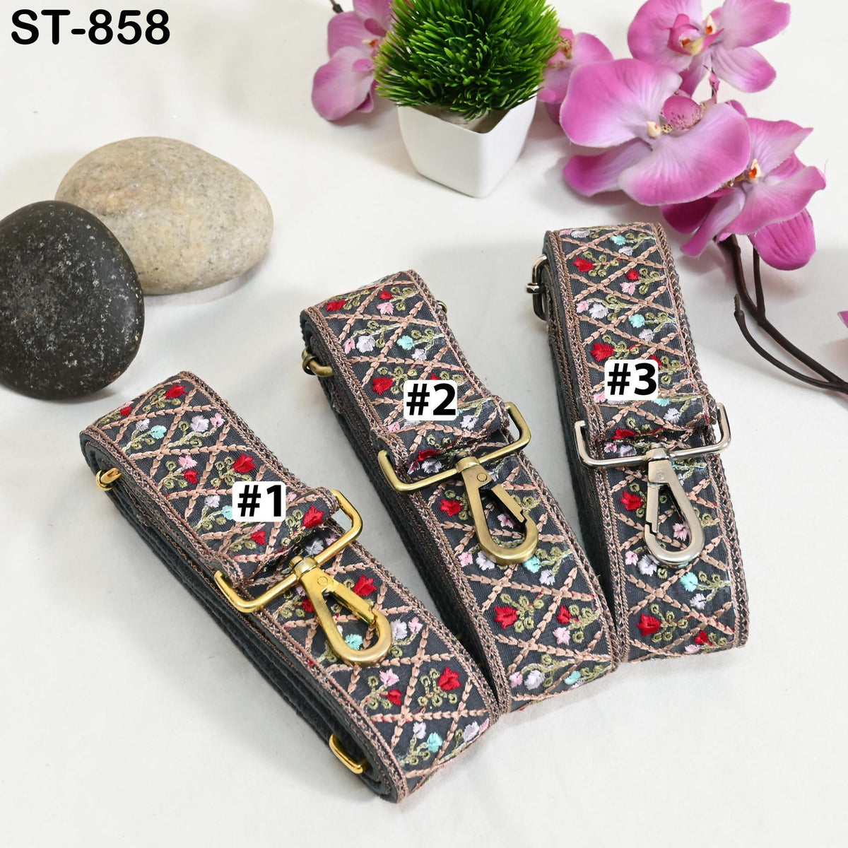 three colorful straps with floral patterns, each with a different number printed on it, placed on a white surface alongside some rocks and pink flowers.