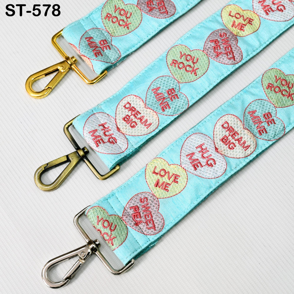 a blue strap with heart-shaped embroidered phrases such as "You Rock", "Be Mine", "Dream Big", "HUG ME", and "Love Me". The strap has a gold hook and a silver loop, and the image includes the text "ST-579" in the top left corner.