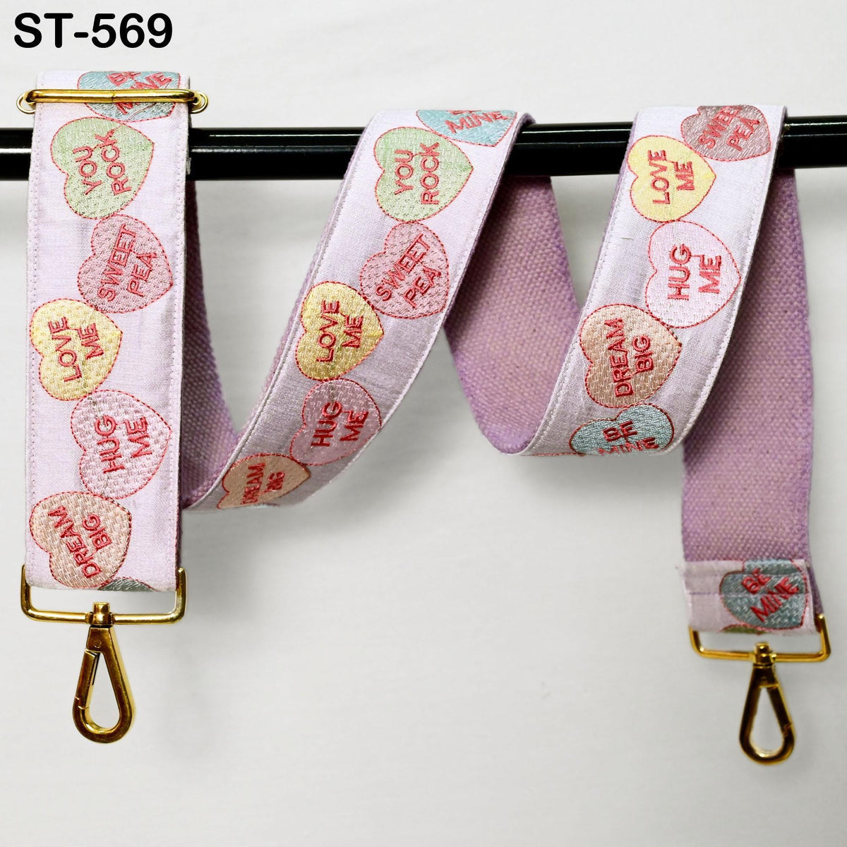 A pink and white heart-shaped strap with various romantic sayings printed on it, hanging from a black hook.