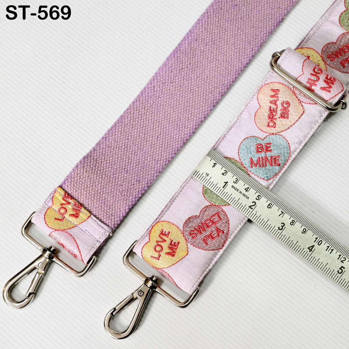 a pink strap with heart-shaped embroidered phrases such as "LOVE ME", "SWEET PEA", and "DREAM BIG". The strap is attached to a metal clasp, and there is a ruler placed next to it, indicating the strap's length.